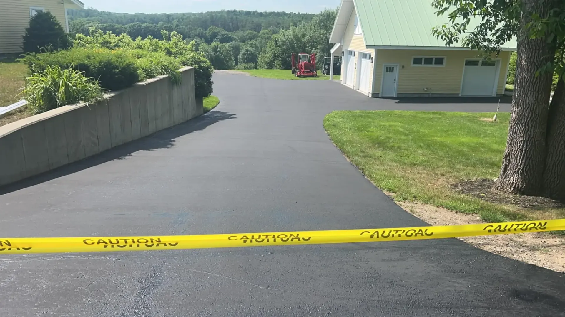 Newly paved black asphalt driveway with caution tape. A yellow house with white garage doors is on the right.