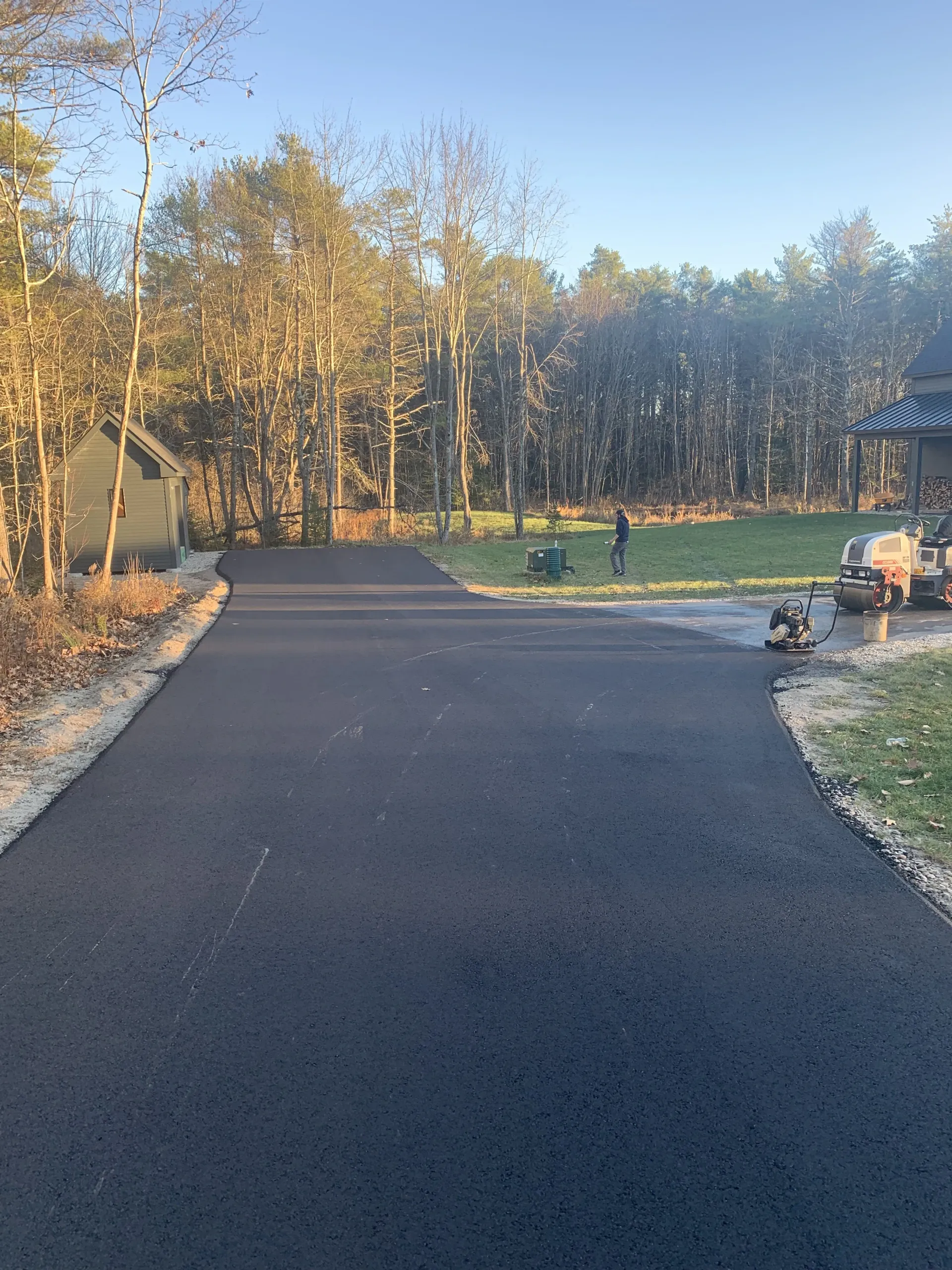 Newly paved black asphalt driveway leading towards trees and a house. Sunlight on the fresh pavement.
