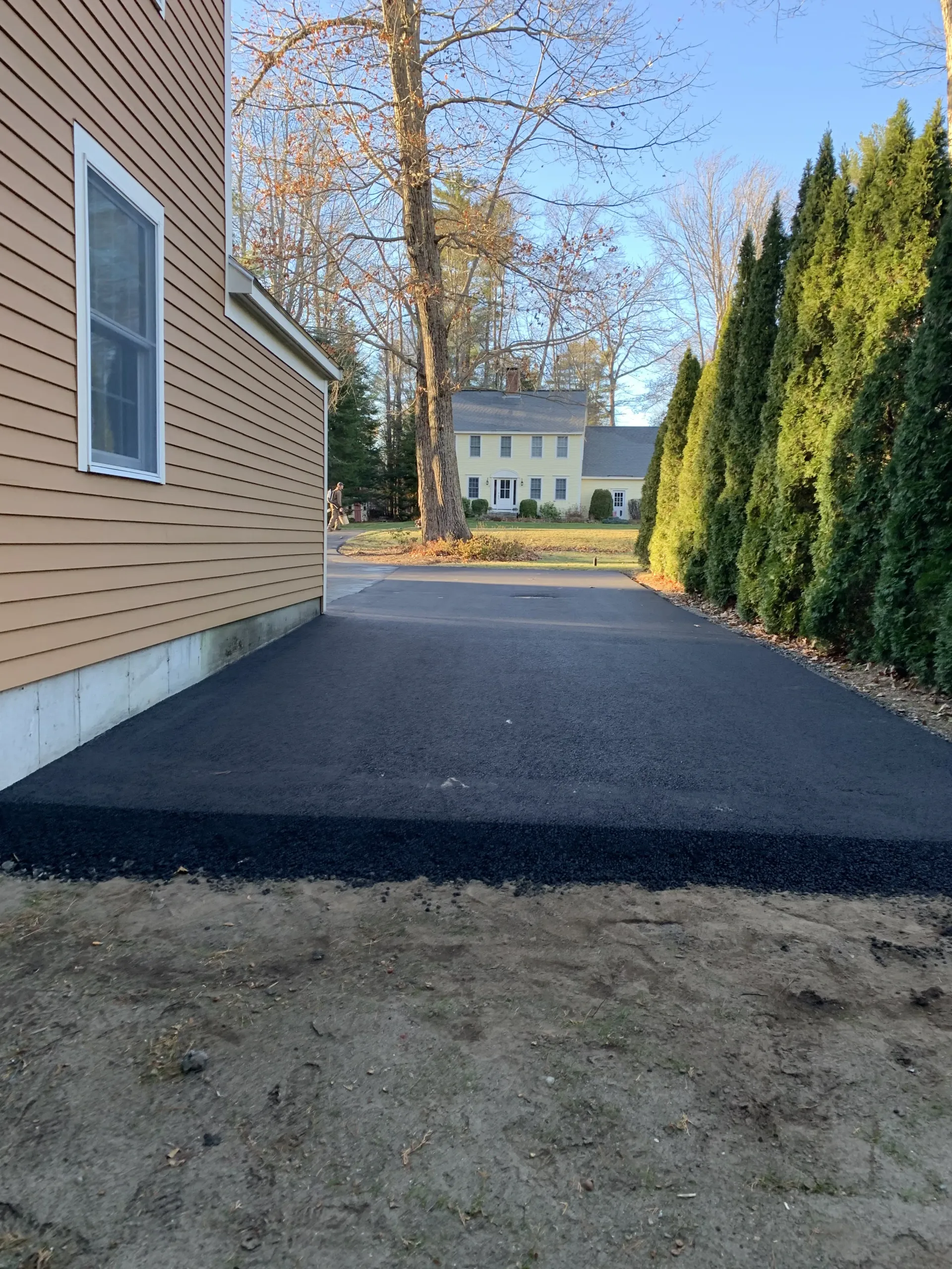 Newly paved black asphalt driveway beside a light brown house and a green hedge.