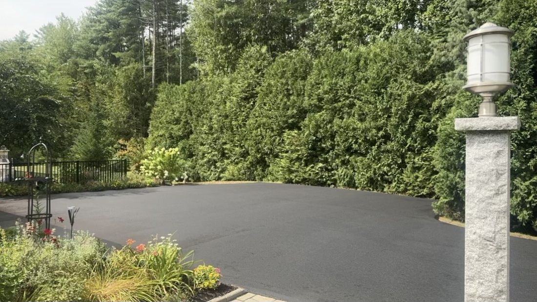 Empty paved driveway bordered by lush green hedges and trees, with a stone lamp post in the foreground