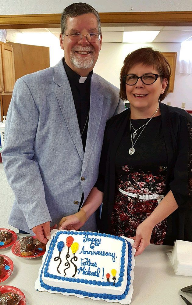 A person in a clerical collar and another person stand behind a cake that reads,