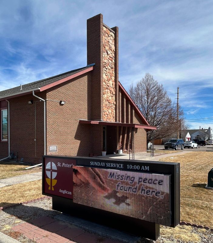 A brick church building with a tall textured chimney and a front sign displaying service times under a blue, cloudy sky.