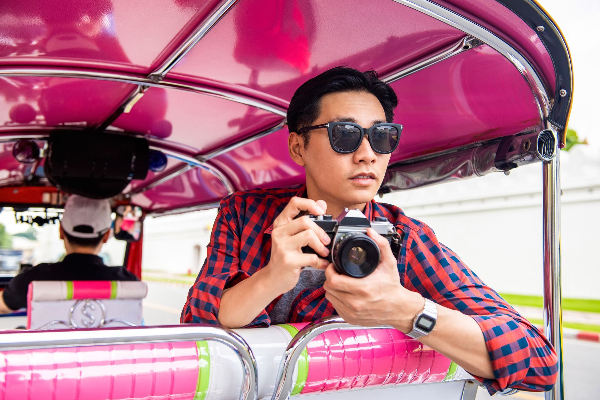 A man in sunglasses takes photos from a pink tuk-tuk. He wears a plaid shirt and a watch, with the city in the background.