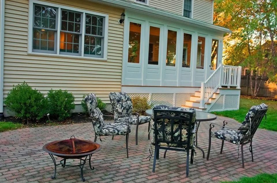 A patio with four patterned chairs, a small round table, and a circular fire pit outside a house with a sunroom.