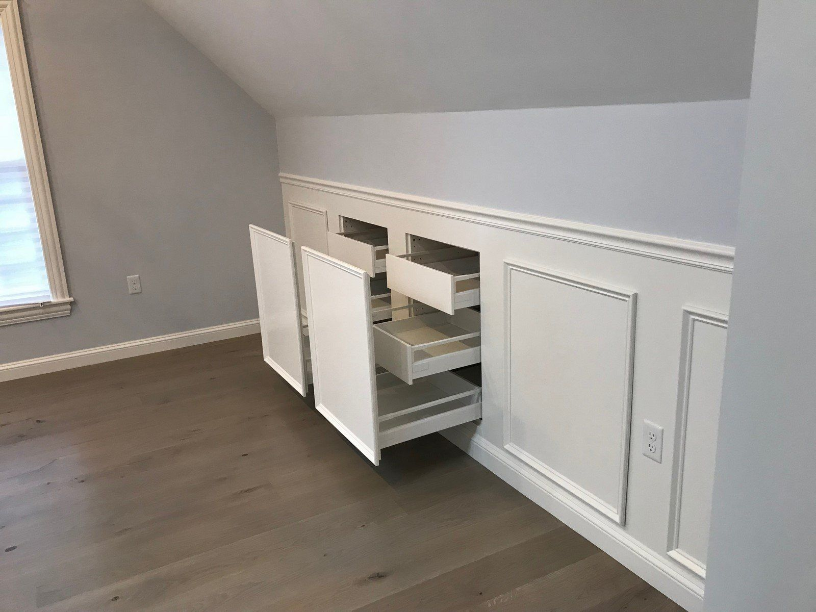 White built-in wall cabinets under a sloped ceiling, with several drawers open to reveal storage in a modern room.