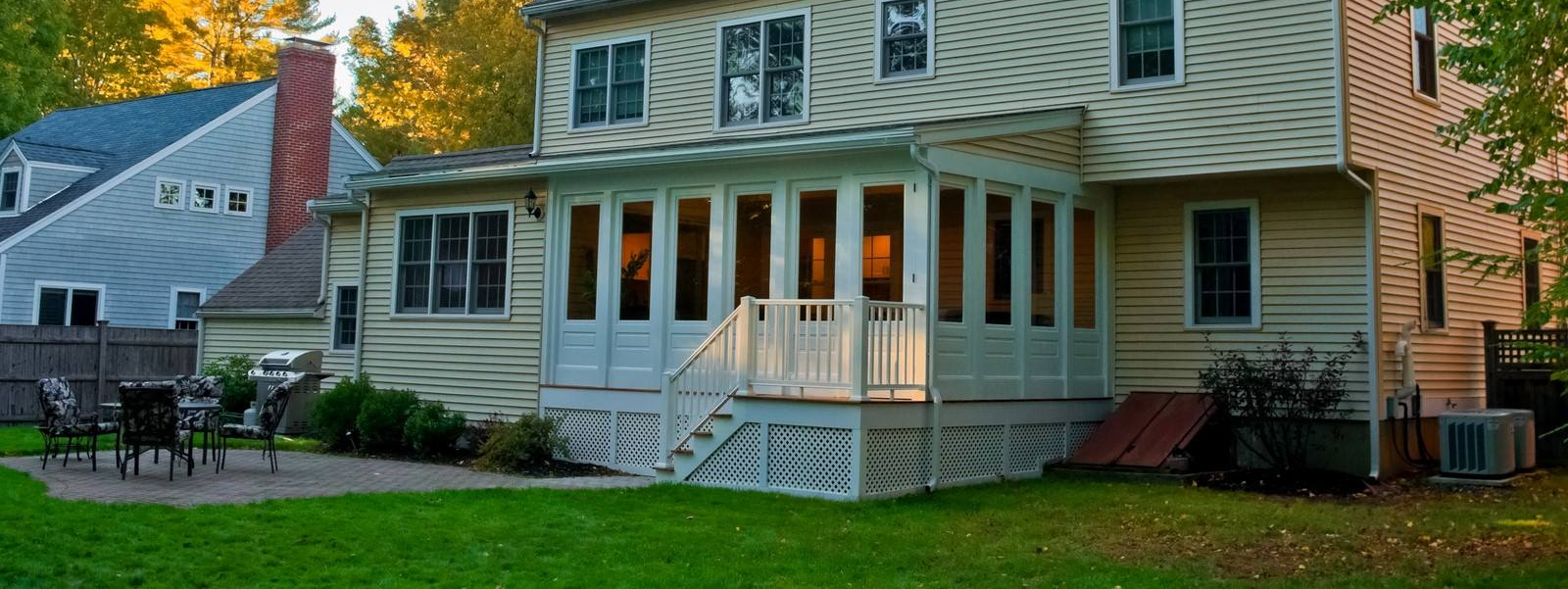 A multi-story house with a white sunroom, a deck, a stone patio with outdoor furniture, and a grassy backyard.