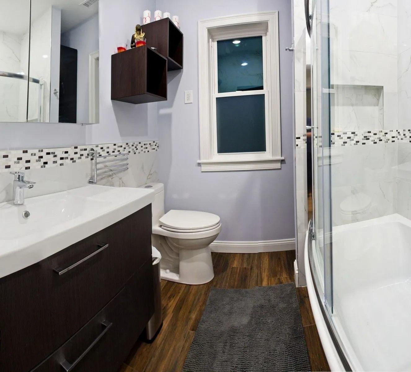 A bathroom with a dark wood vanity, toilet, and glass-enclosed shower, featuring light purple walls and wood flooring.