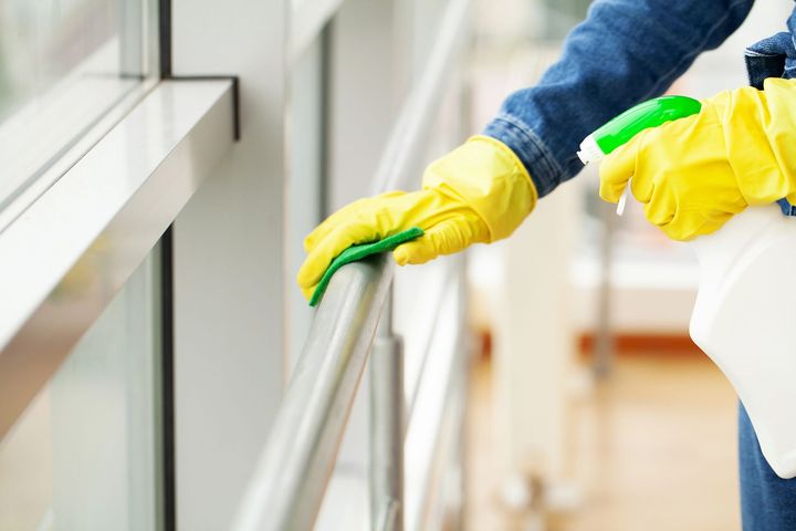 Una persona con i guanti gialli pulisce una finestra con uno spruzzino verde e un panno bianco.