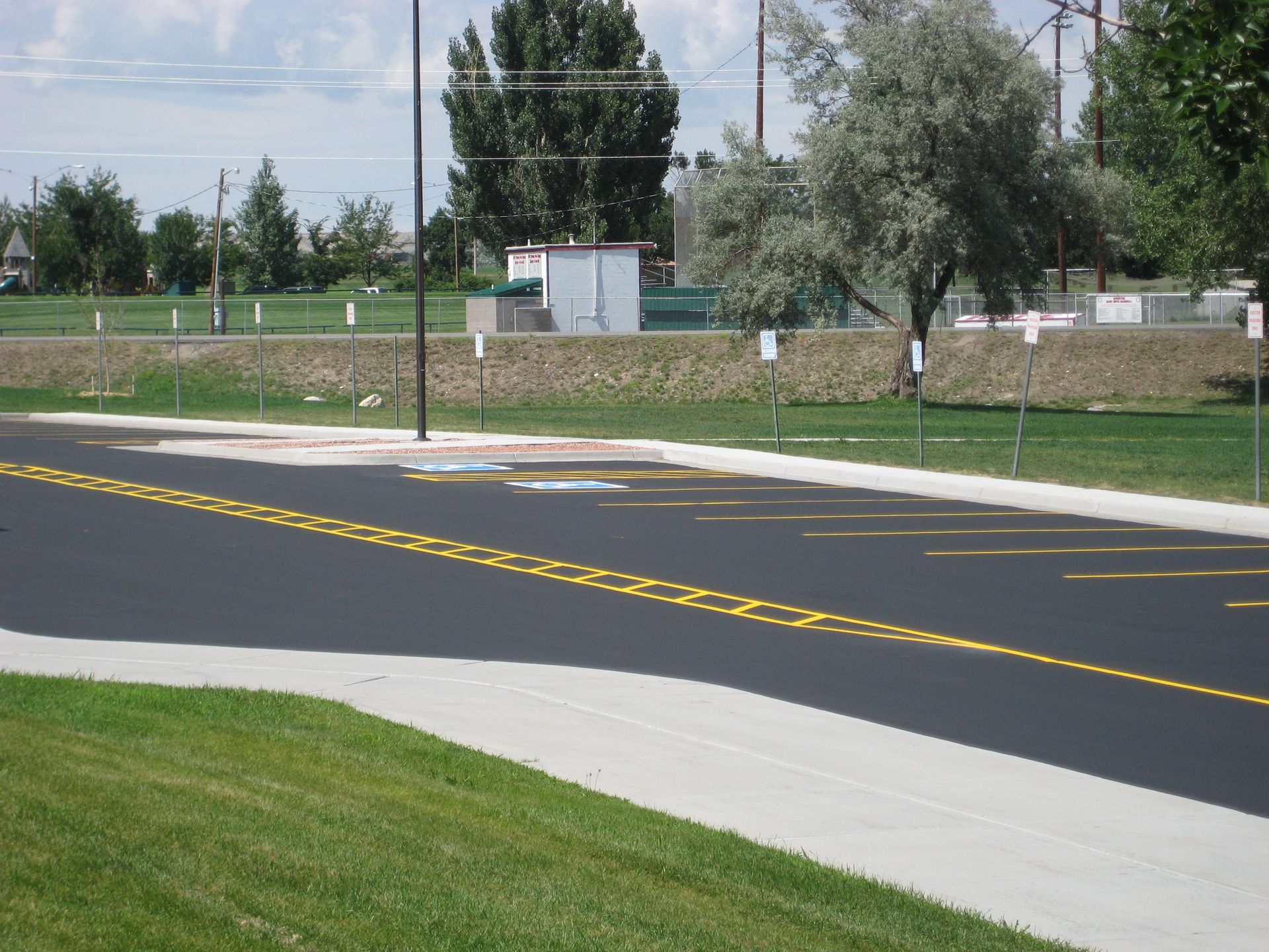 Freshly paved road with double yellow lines beside a sidewalk, grass, and trees on a sunny day