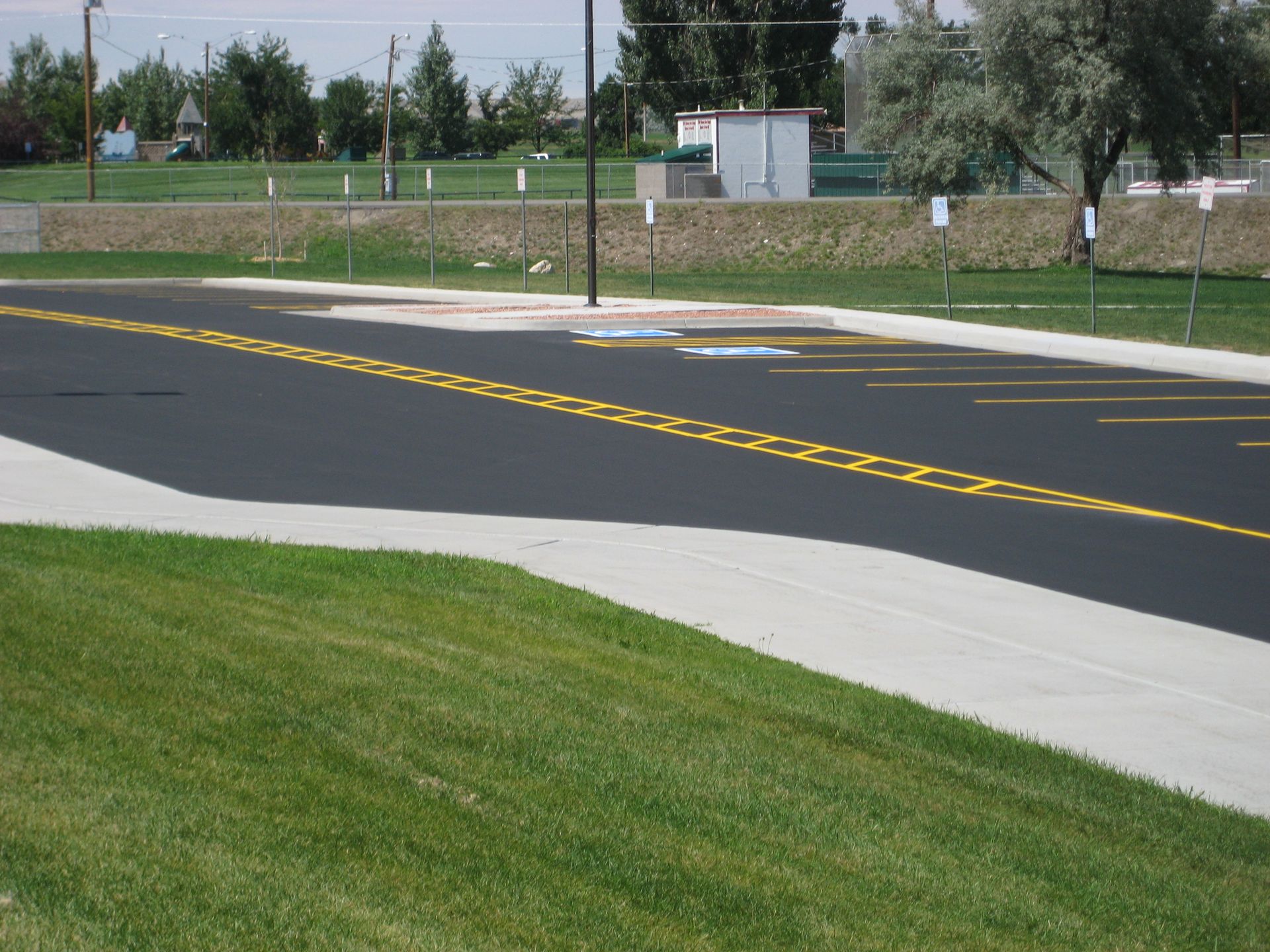 Curved paved road with yellow center lines beside a grassy slope and sidewalk near trees and a fence