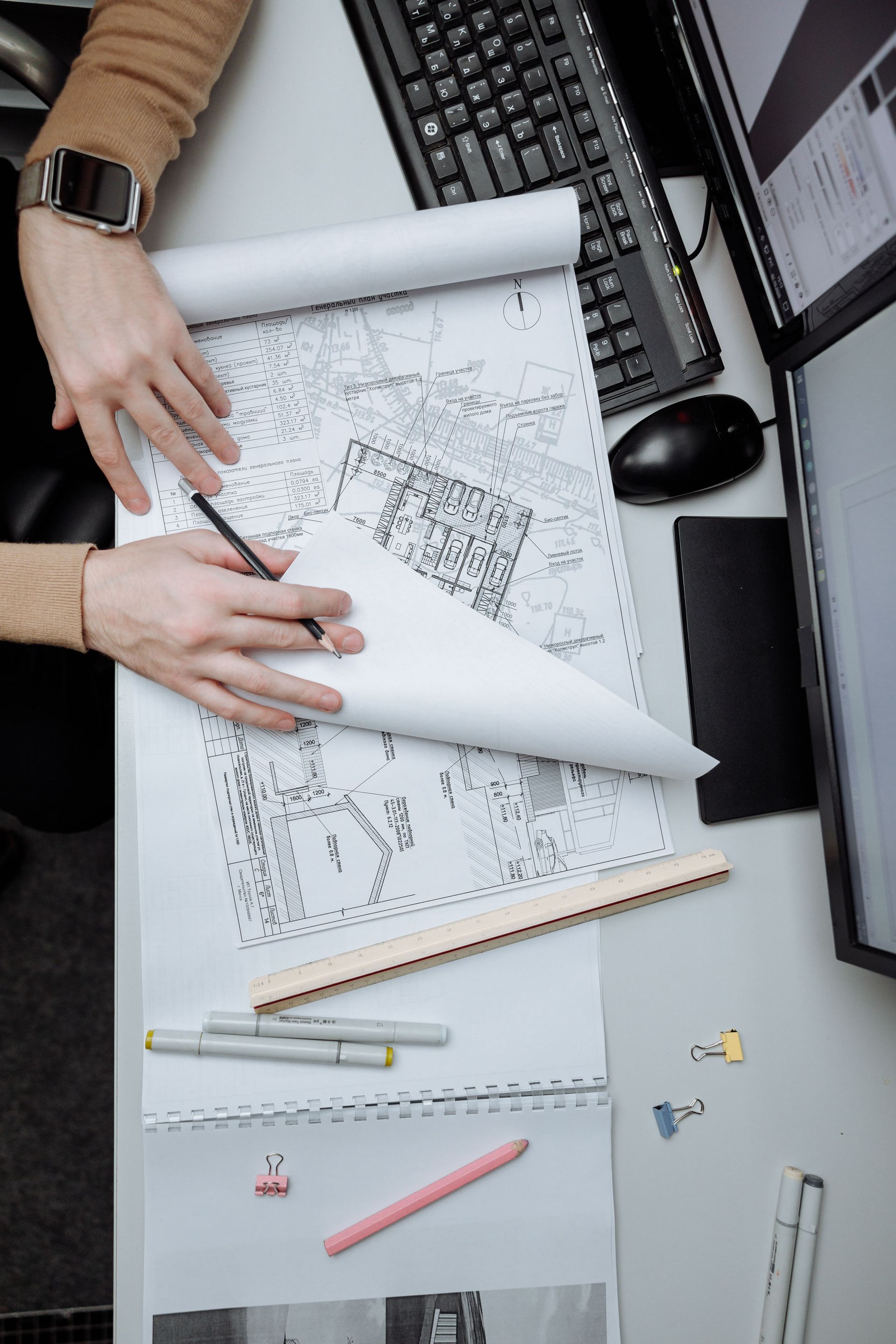 Hands sketching architectural plans on a desk with ruler, laptop, and scattered pencils