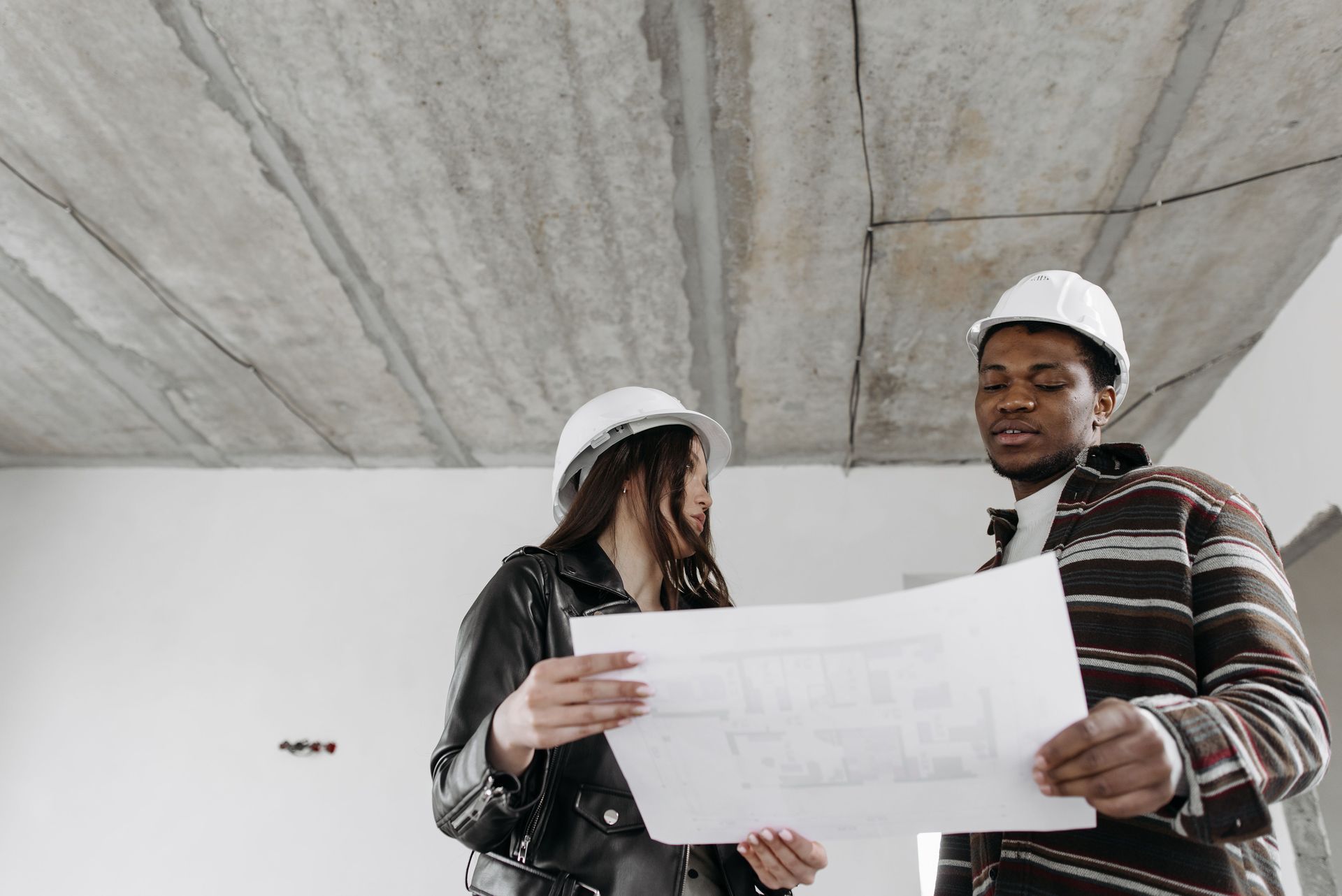 Two construction workers in hard hats reviewing blueprints under a concrete overhang