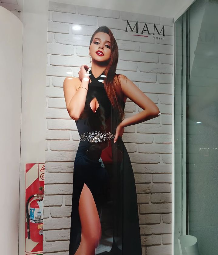 Una mujer con un vestido de noche negro posa frente a una pared de ladrillos blancos. Se ve el logo de