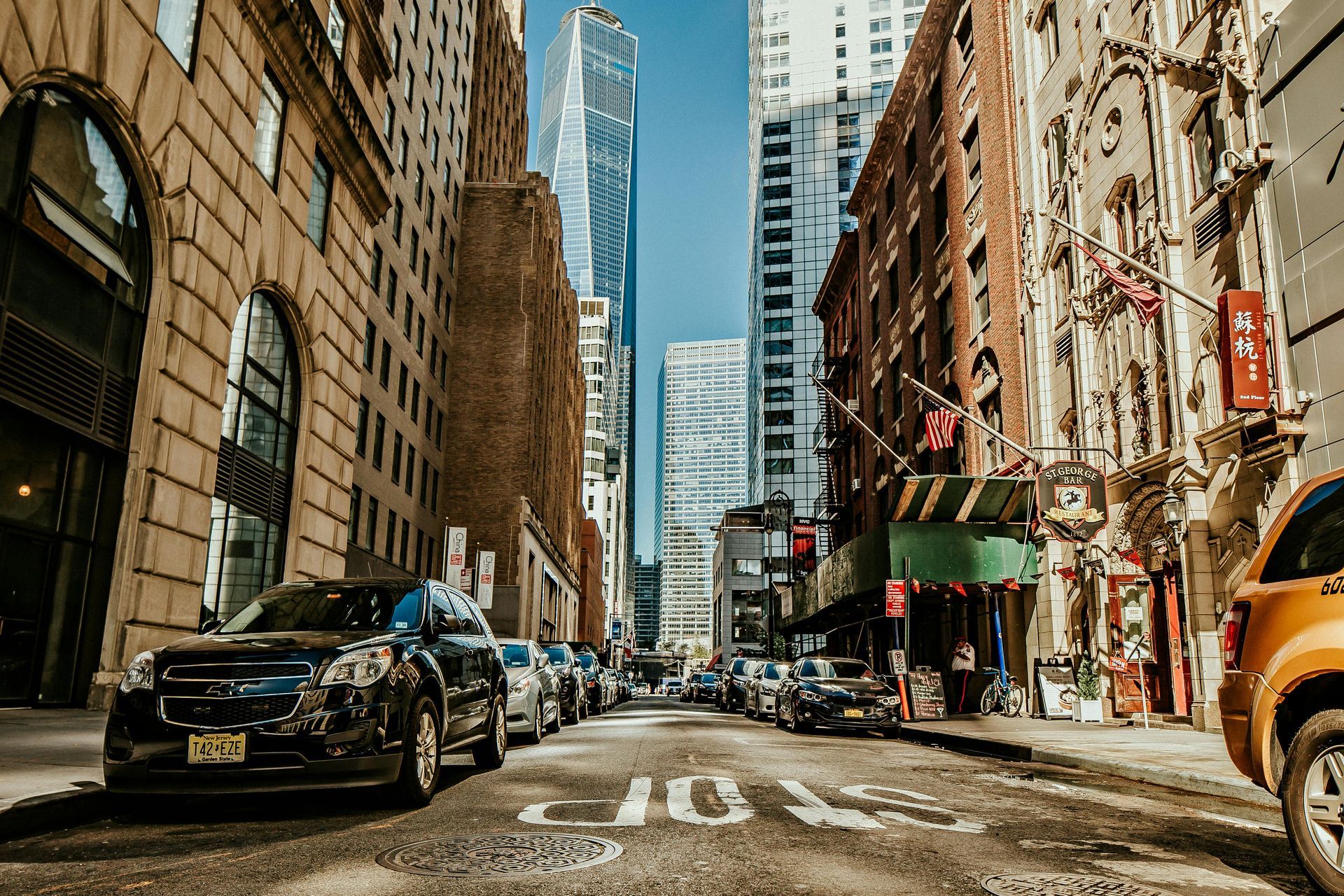 Street in New York City with parked cars, tall buildings, and a clear blue sky.