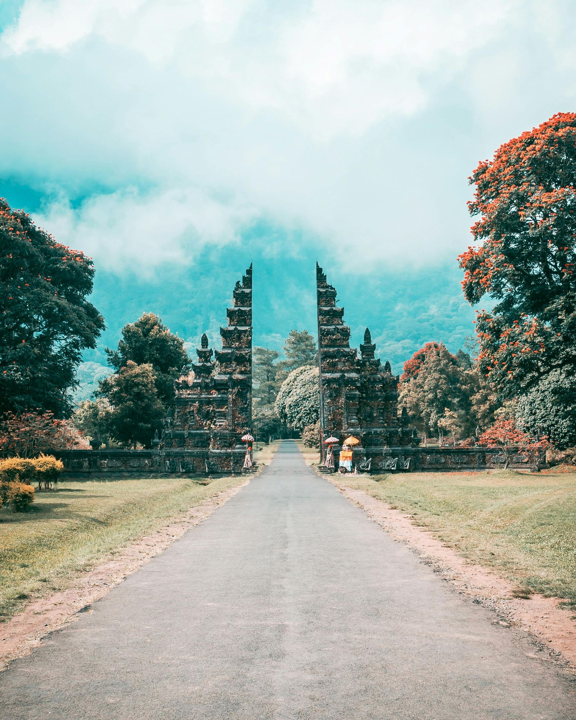 Stone gate with ornate carvings leads to a road in Bali, Indonesia; trees and clouds in the background.