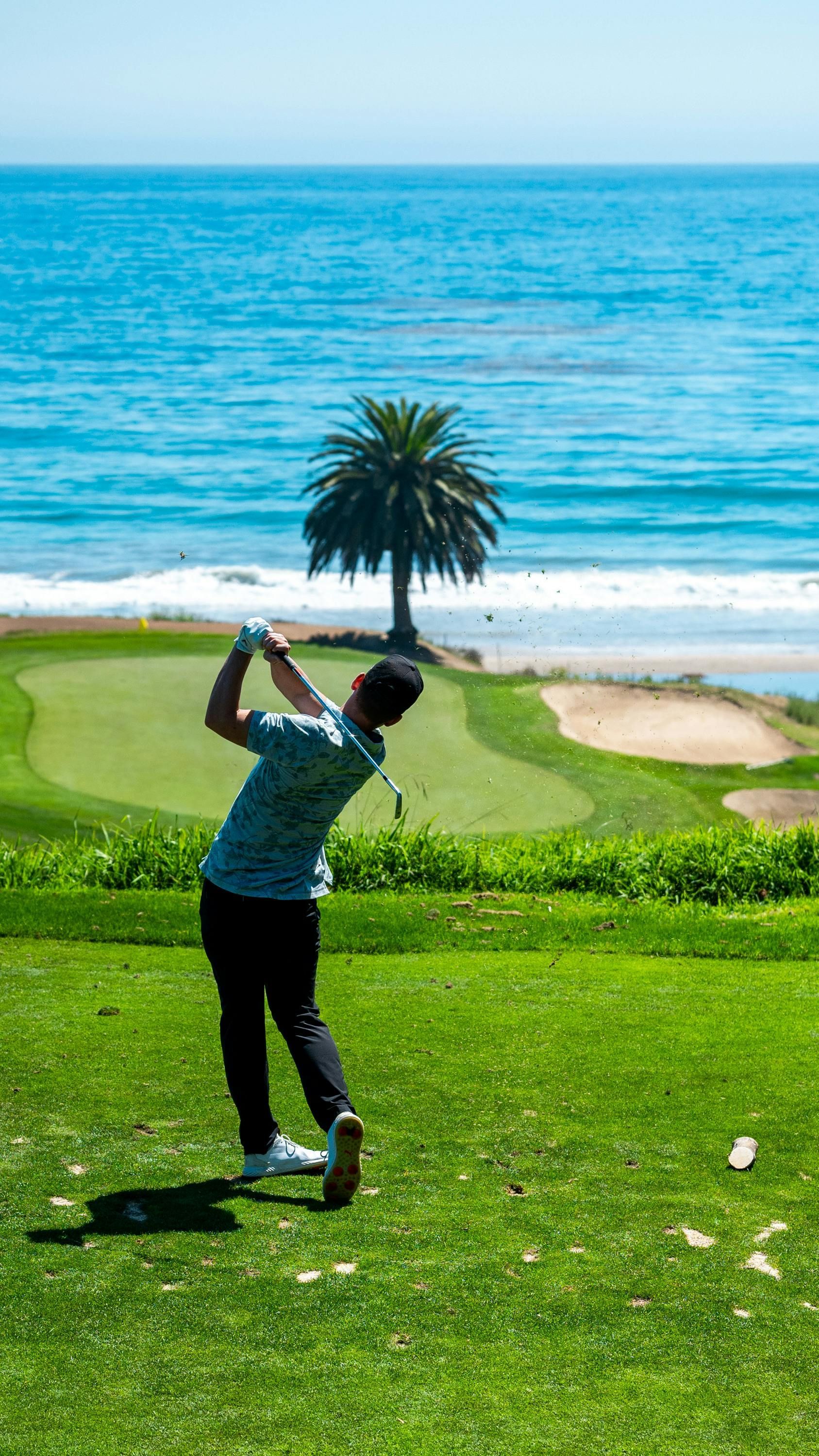 Golfer in motion, teeing off on a green golf course near the ocean. Sunny day, blue water, palm tree.
