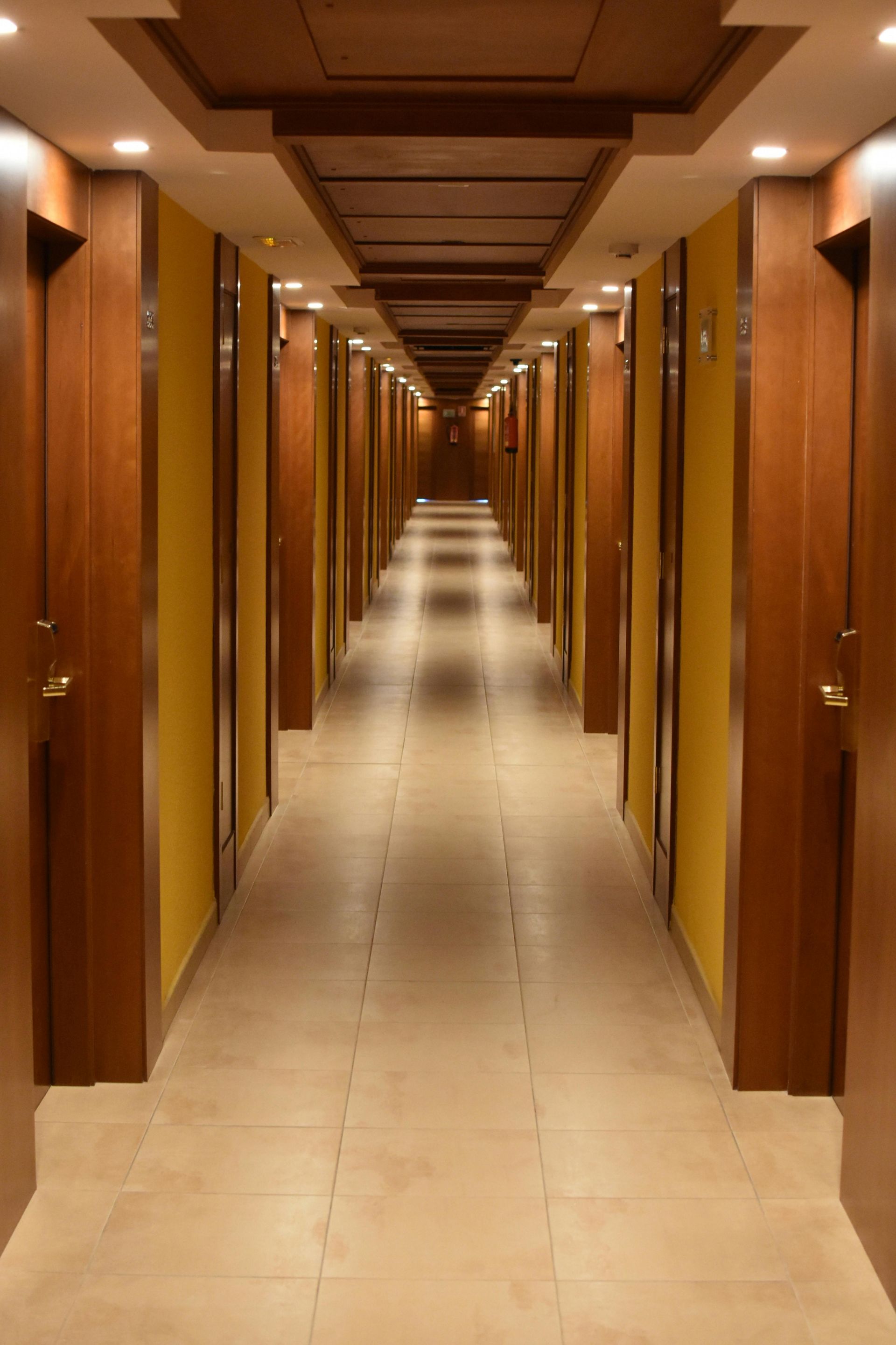Long hotel hallway with wooden doors and ceiling panels, yellow walls, and tan tiled floor.