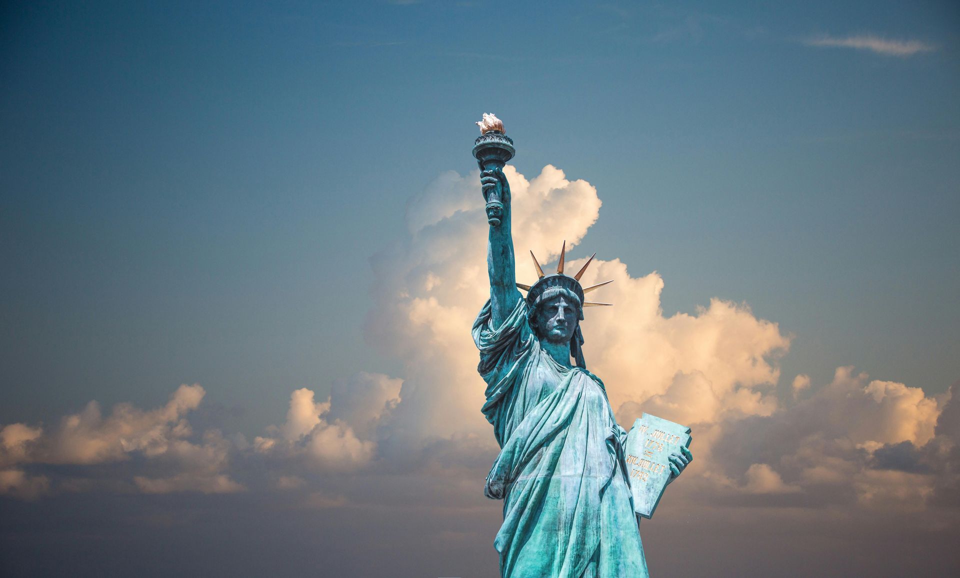 Statue of Liberty, green with raised torch, against a partly cloudy sky.