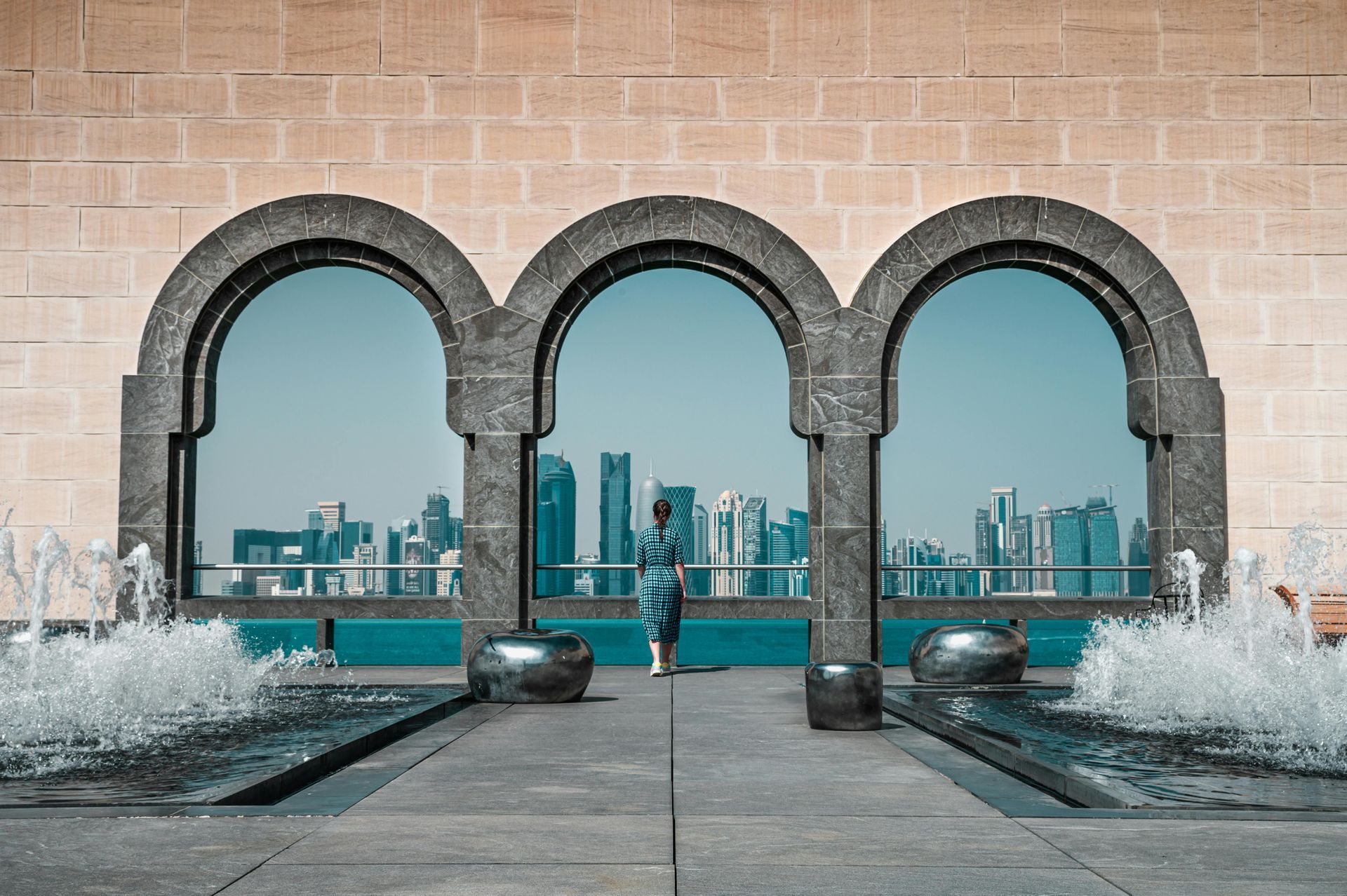 Person standing in arched windows, looking at Doha skyline. Fountains flank the walkway, beige building.
