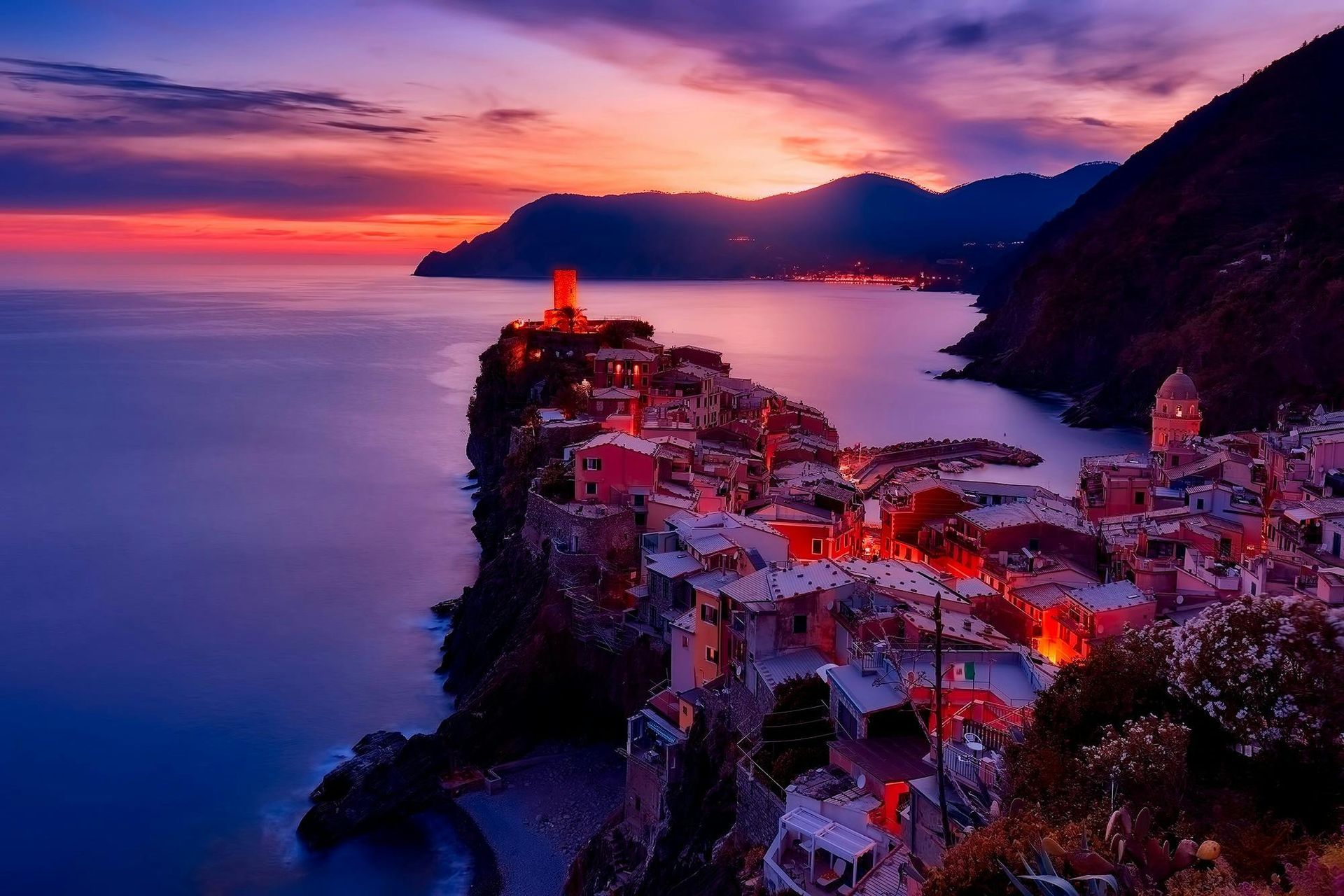 Coastal Italian village at dusk, colorful buildings perched on cliffs, reflecting lights on water under a vibrant sunset.
