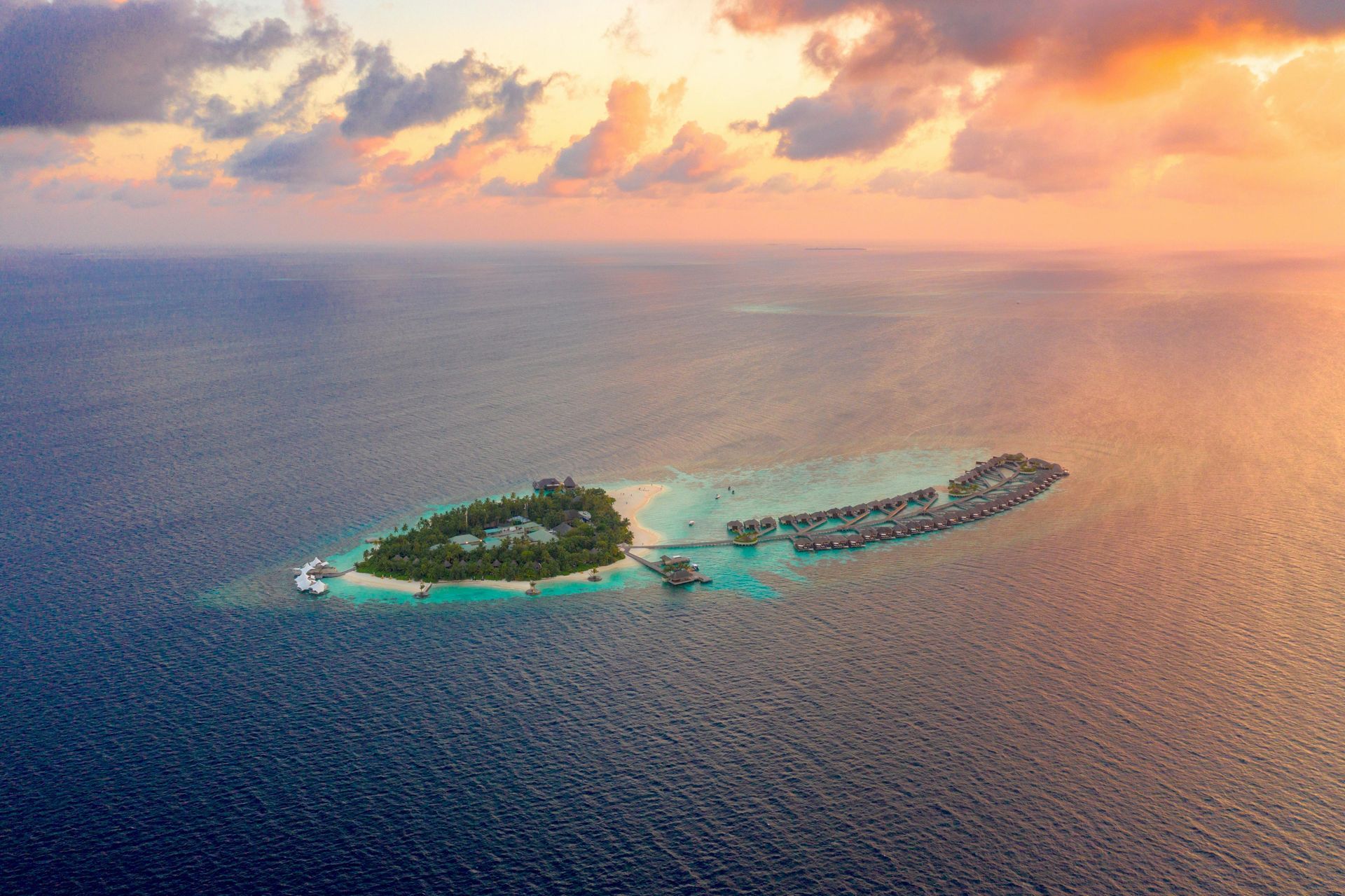 Aerial view of a tropical island with overwater bungalows, surrounded by turquoise water, under a sunset sky.