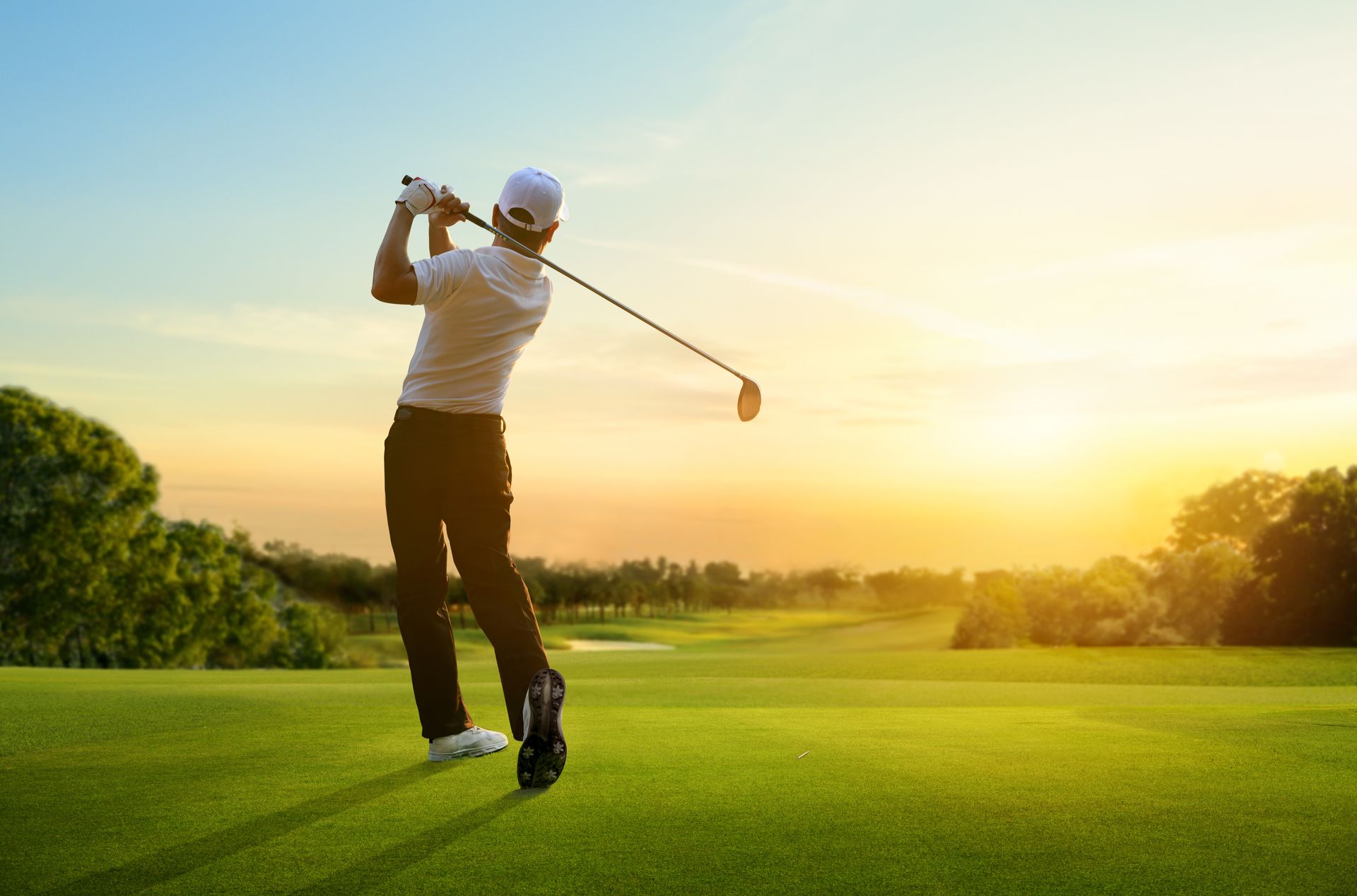 Golfer swinging a club on a green course, sunset in the background.