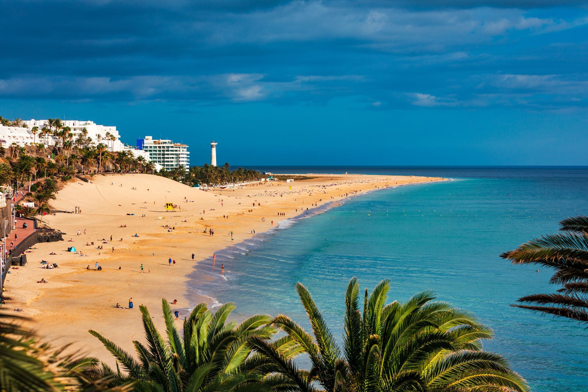 Sandy beach and turquoise water under a blue sky, palm trees in the foreground, white buildings and a lighthouse.