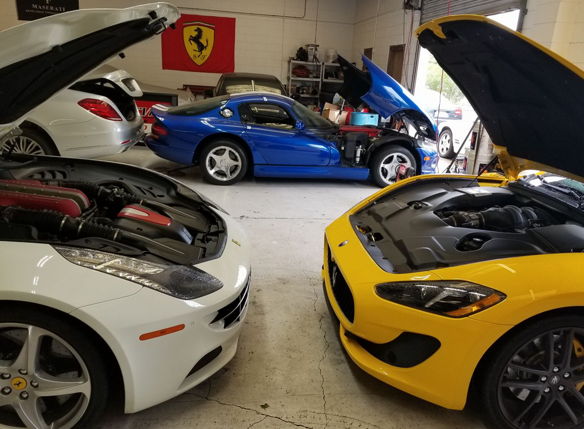 Three sports cars are parked in a garage with their hoods open.