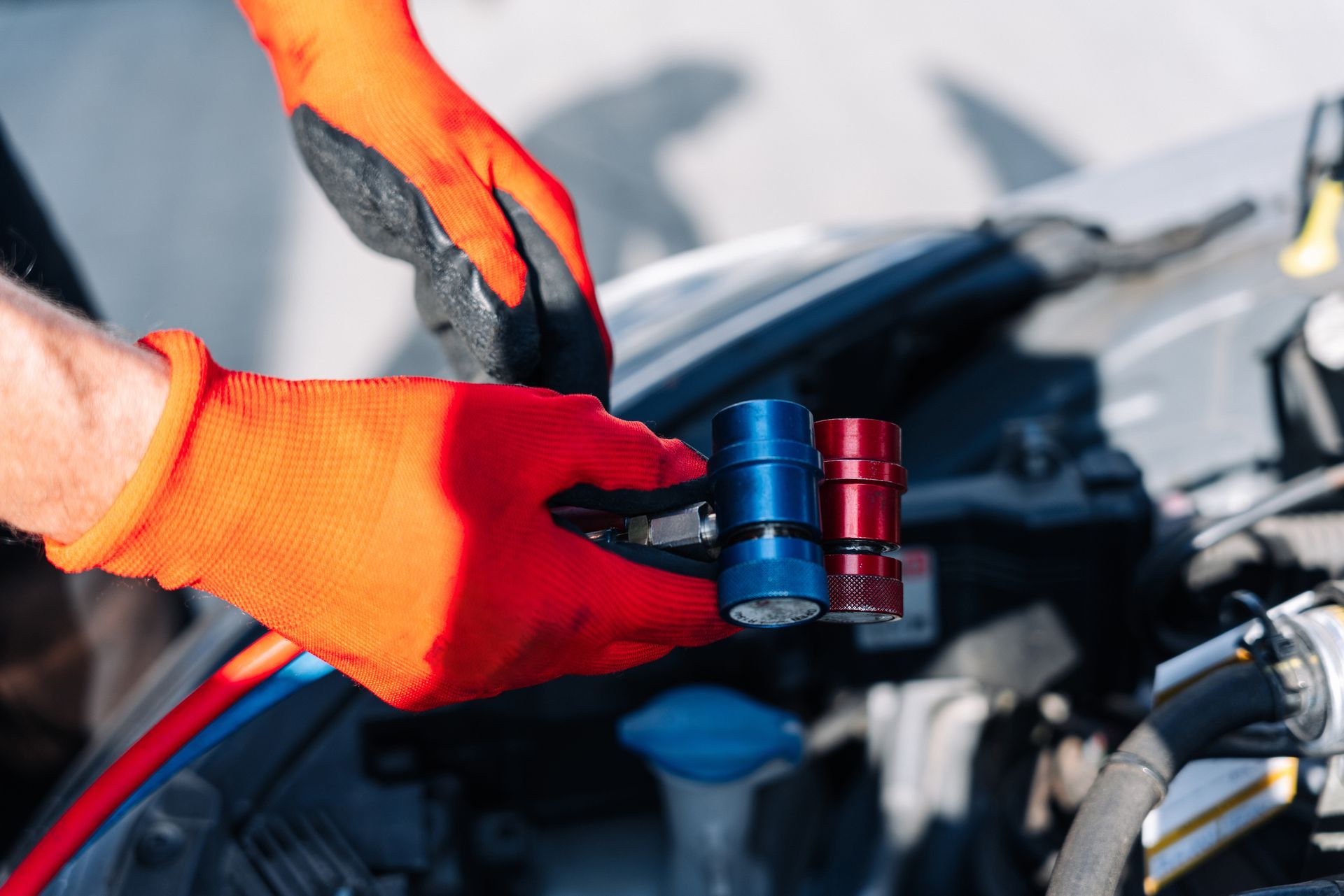 Des mains gantées d'orange tiennent des coupleurs de service de climatisation bleus et rouges près d'un moteur de voiture.
