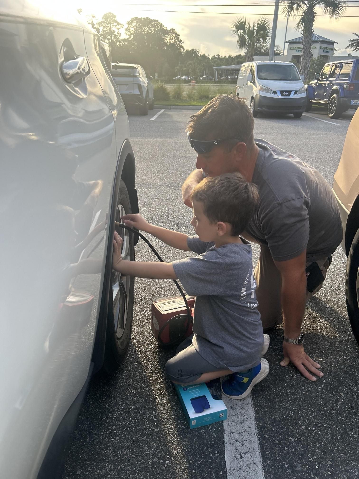A man and a child inflating a car tire in a parking lot. The child holds the air hose while the man watches.