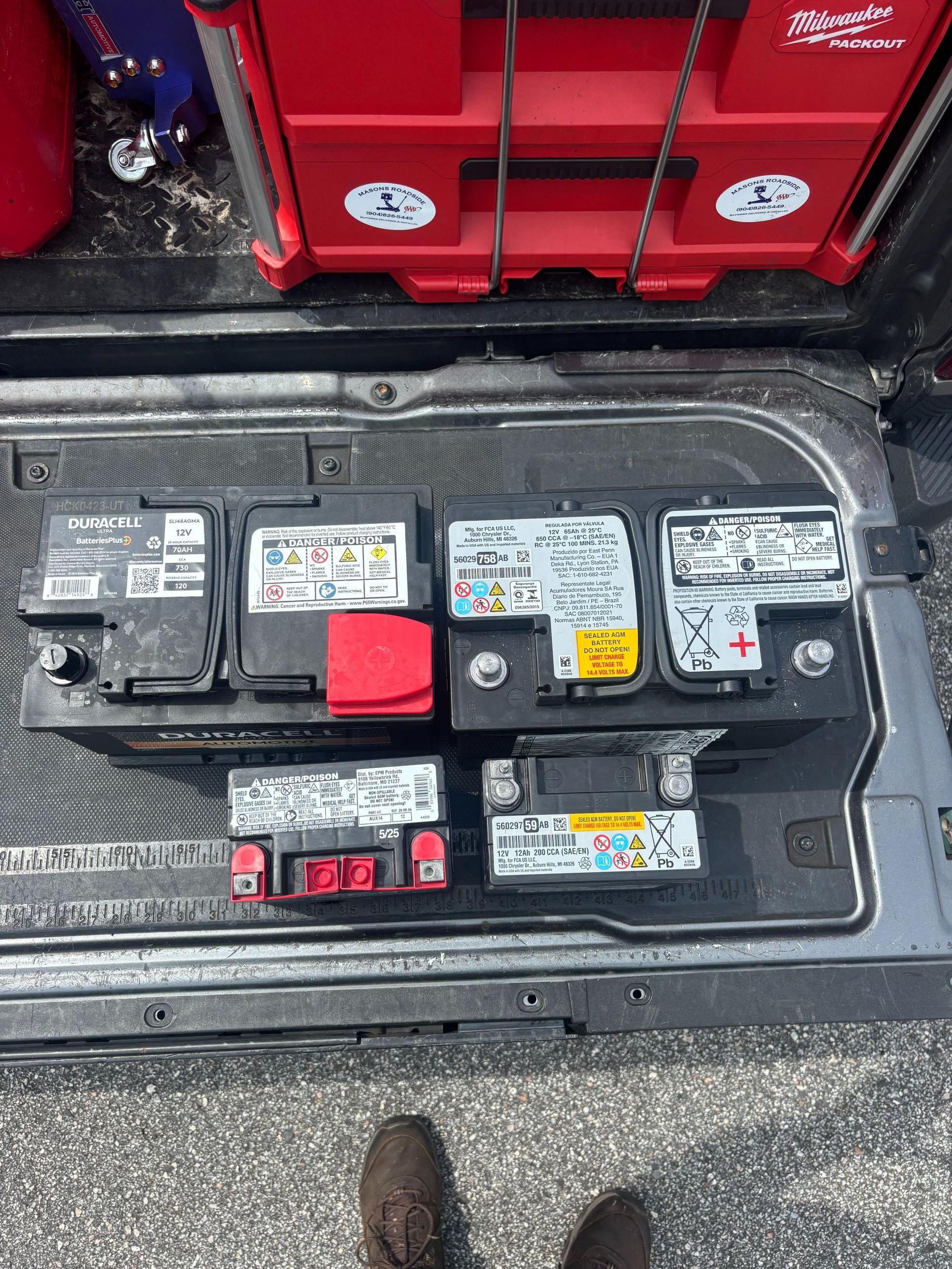 Six car batteries of different sizes sit on a tray inside a vehicle. Red toolbox in background.