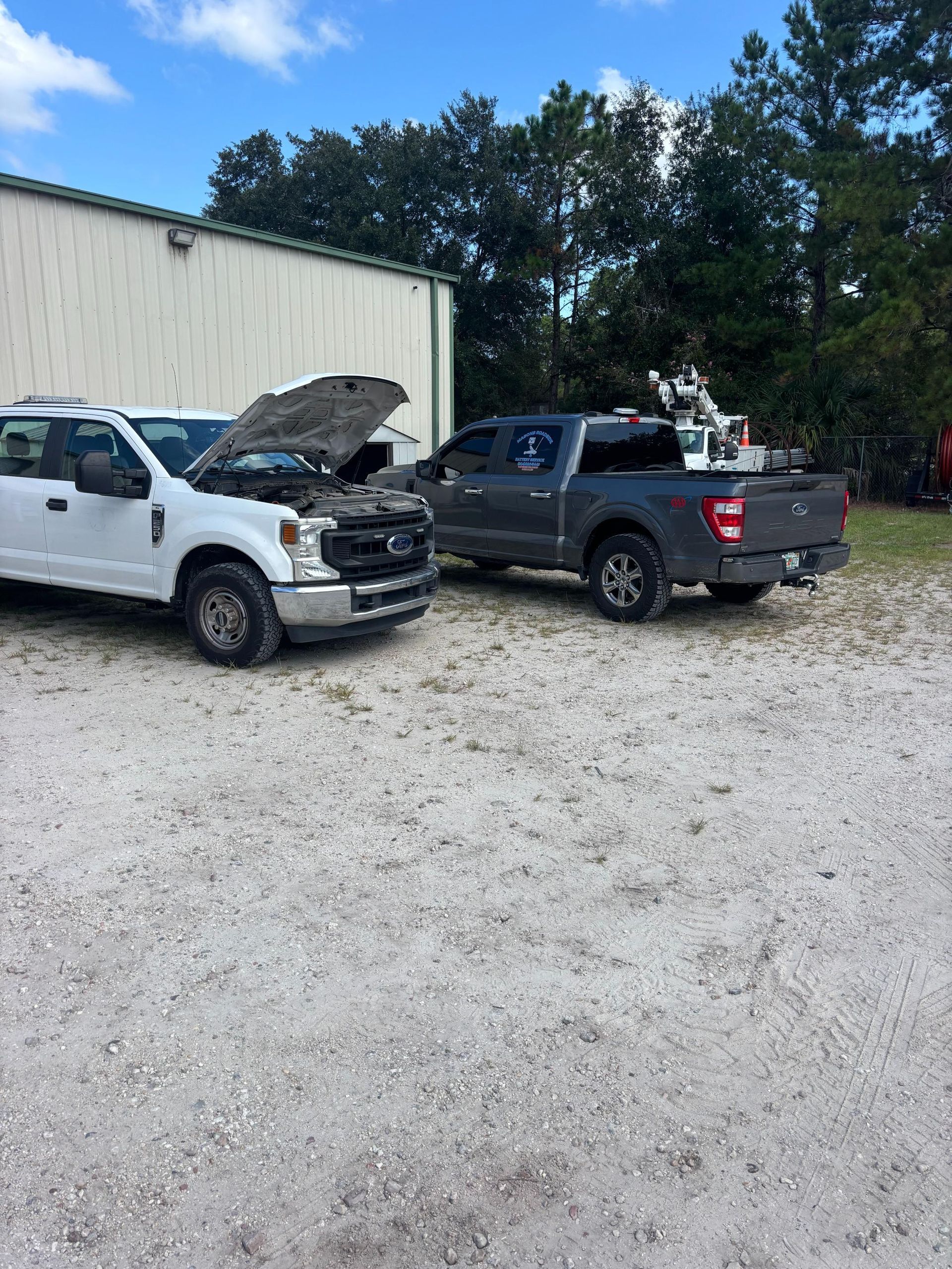 White truck with open hood next to a gray truck parked on gravel, with a building in the background.