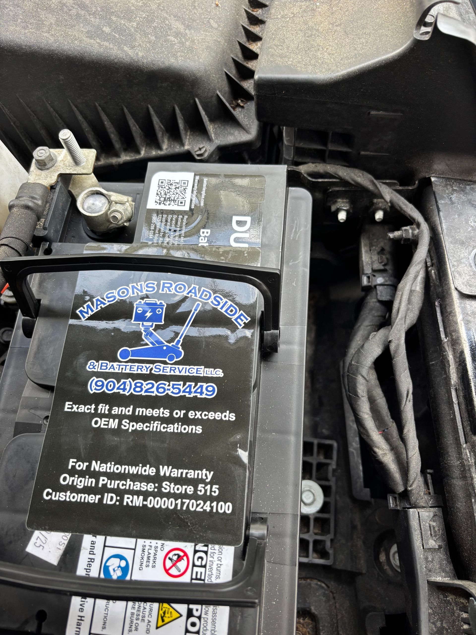Car battery with a sticker from a roadside assistance service, located in an engine bay.