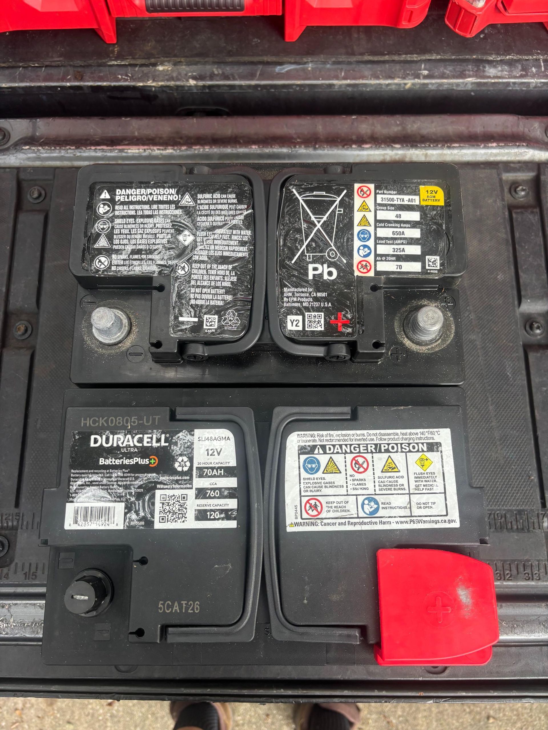 Two car batteries side by side in a tray; the top one is older and the bottom one is new.
