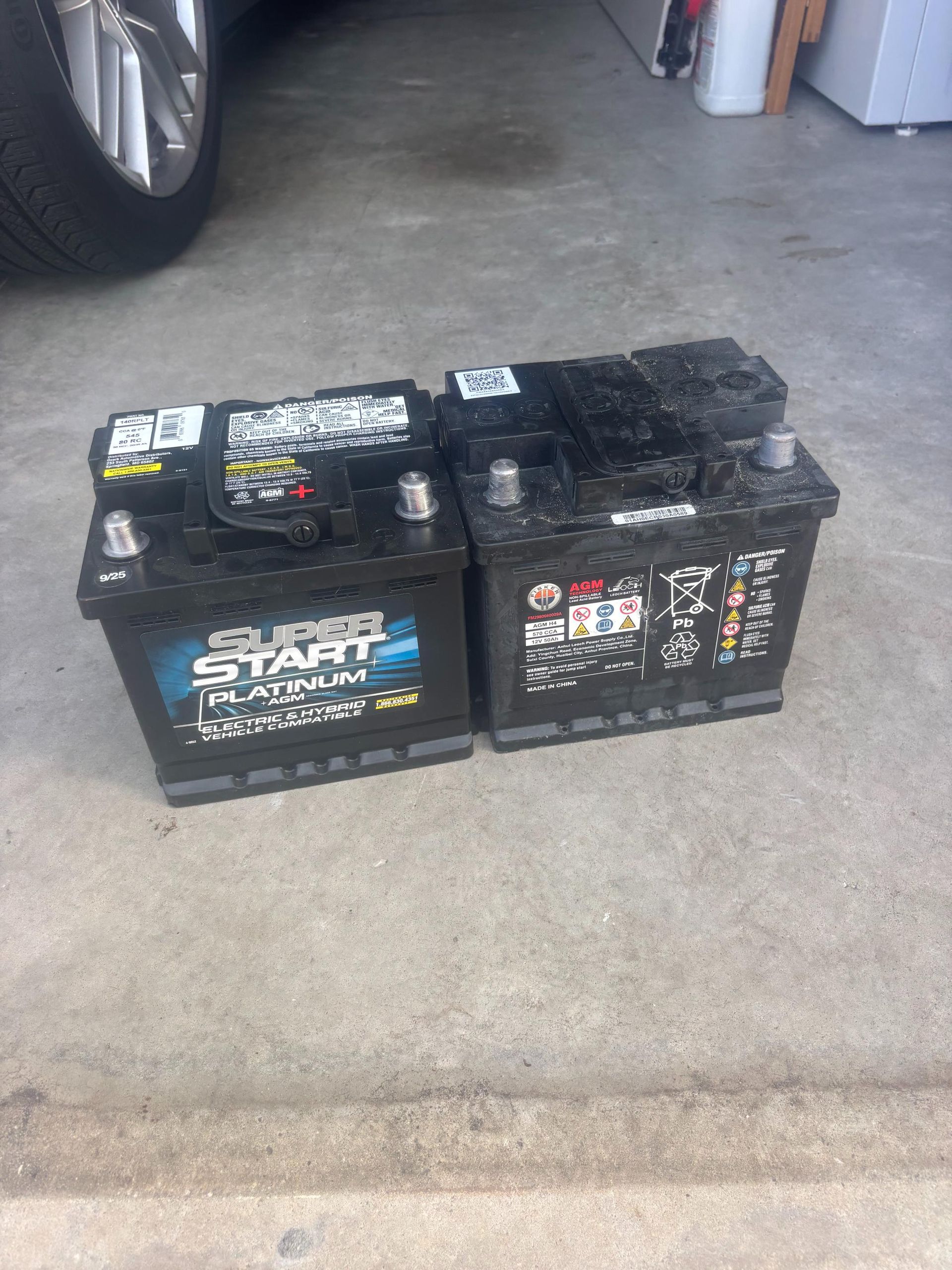 Two black car batteries on a concrete surface. One has a blue label. A car wheel is in the background.