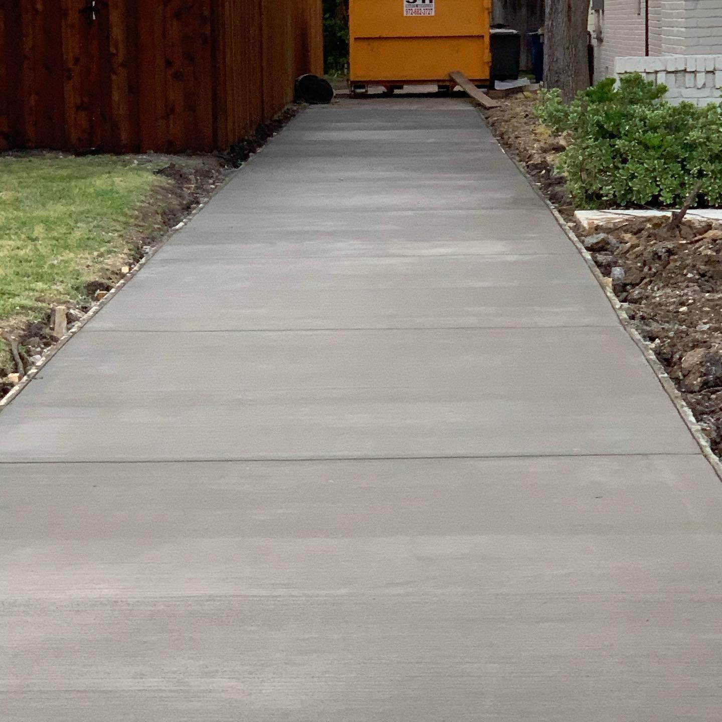Newly poured concrete sidewalk leading towards a yellow dumpster.