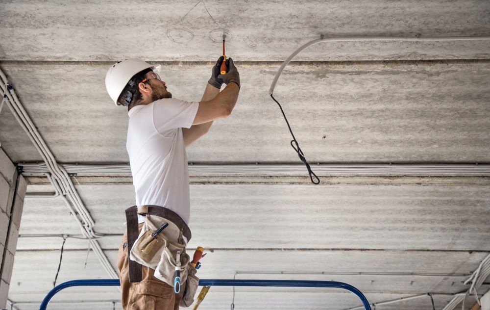 An Electrician Wearing A White Helmet Works On Wiring A Ceiling, Standing On A Ladder — Aliance Group in Bonville, NSW
