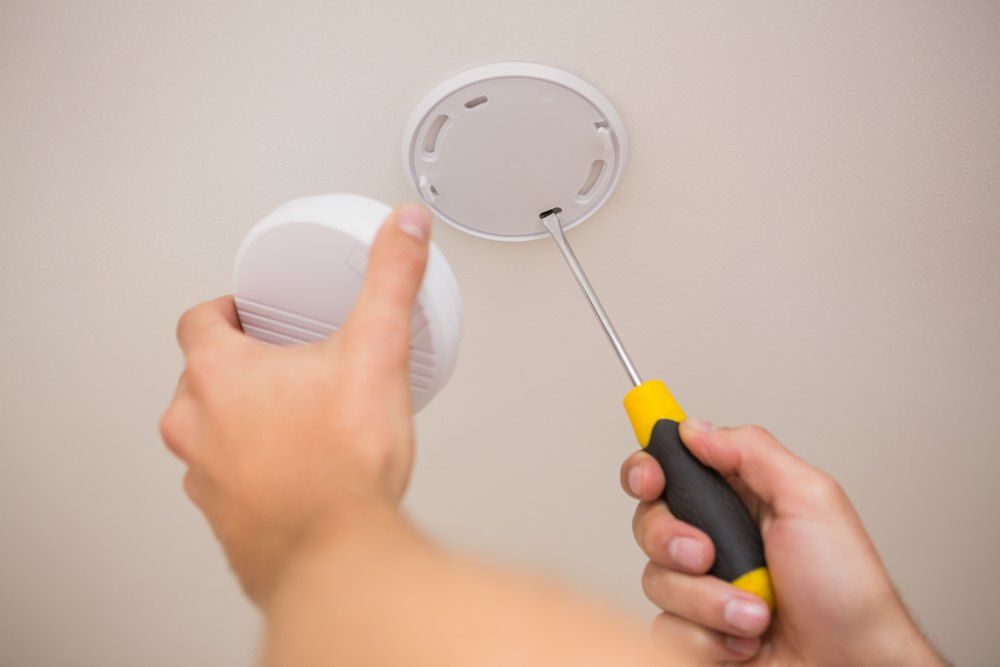 Person Installing A Smoke Detector On A Ceiling Using A Screwdriver — Aliance Group in Kempsey, NSW