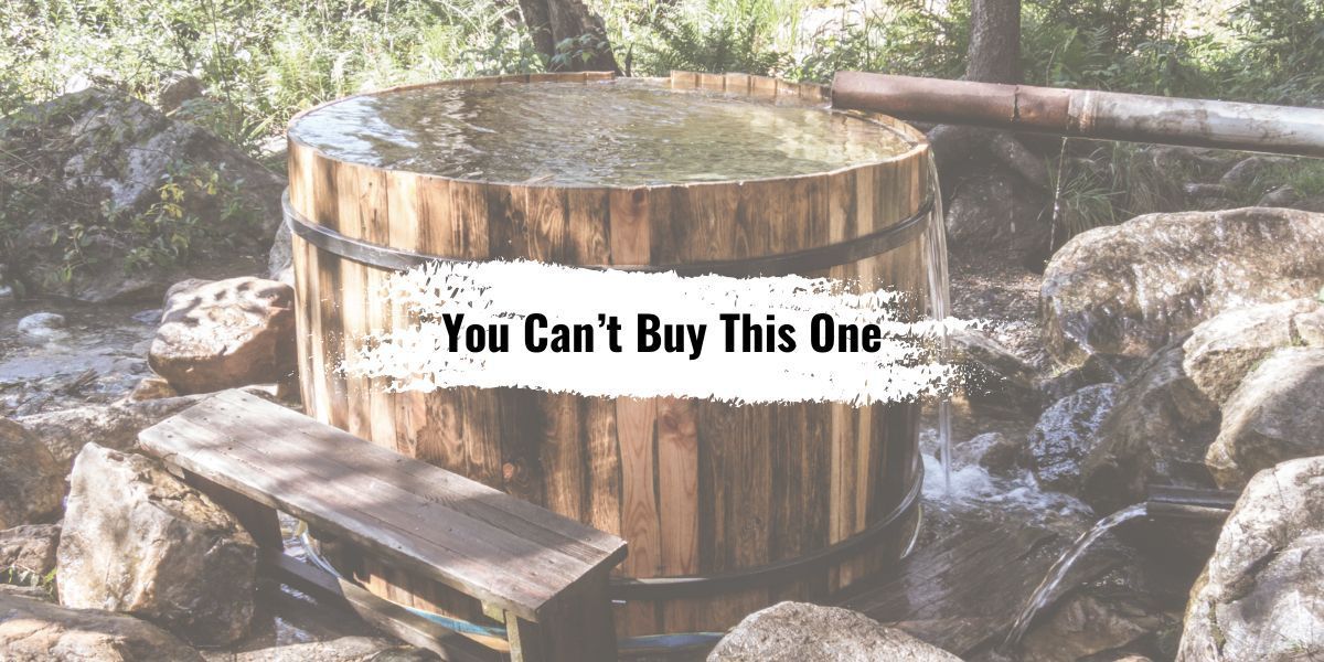 A wooden hot tub in a rustic outdoor setting with a sign reading, “You Can’t Buy This One.”