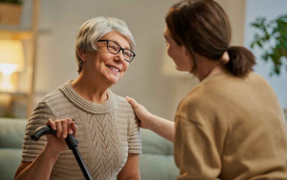 An elderly woman with a cane is sitting on a couch talking to a younger woman.