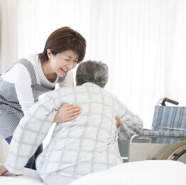 A woman is helping an elderly man in a wheelchair get out of bed.