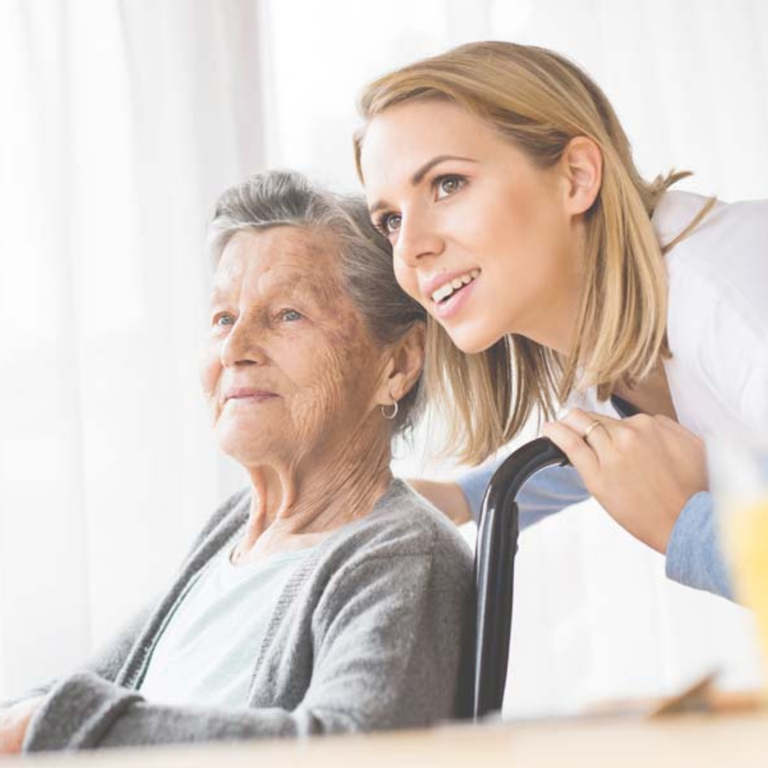 A young woman is standing next to an older woman in a wheelchair.