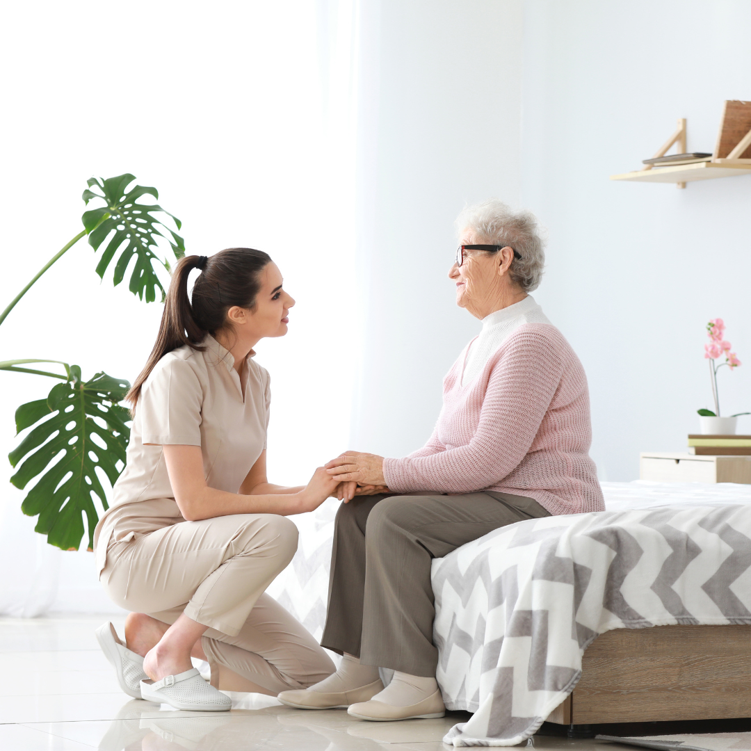 A woman is kneeling down next to an older woman sitting on a bed holding hands.