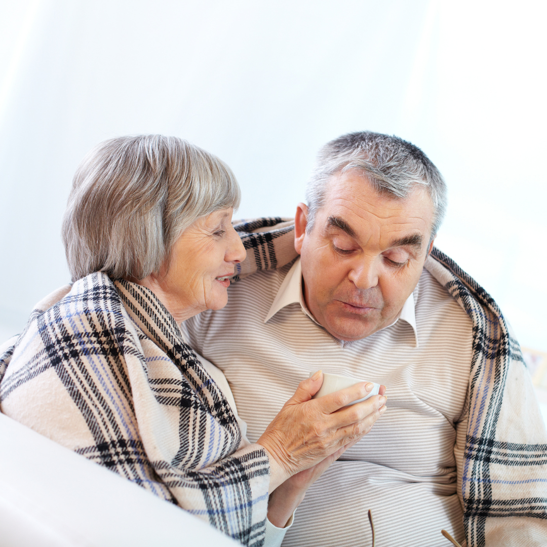 An elderly couple wrapped in a plaid blanket are sitting on a couch.