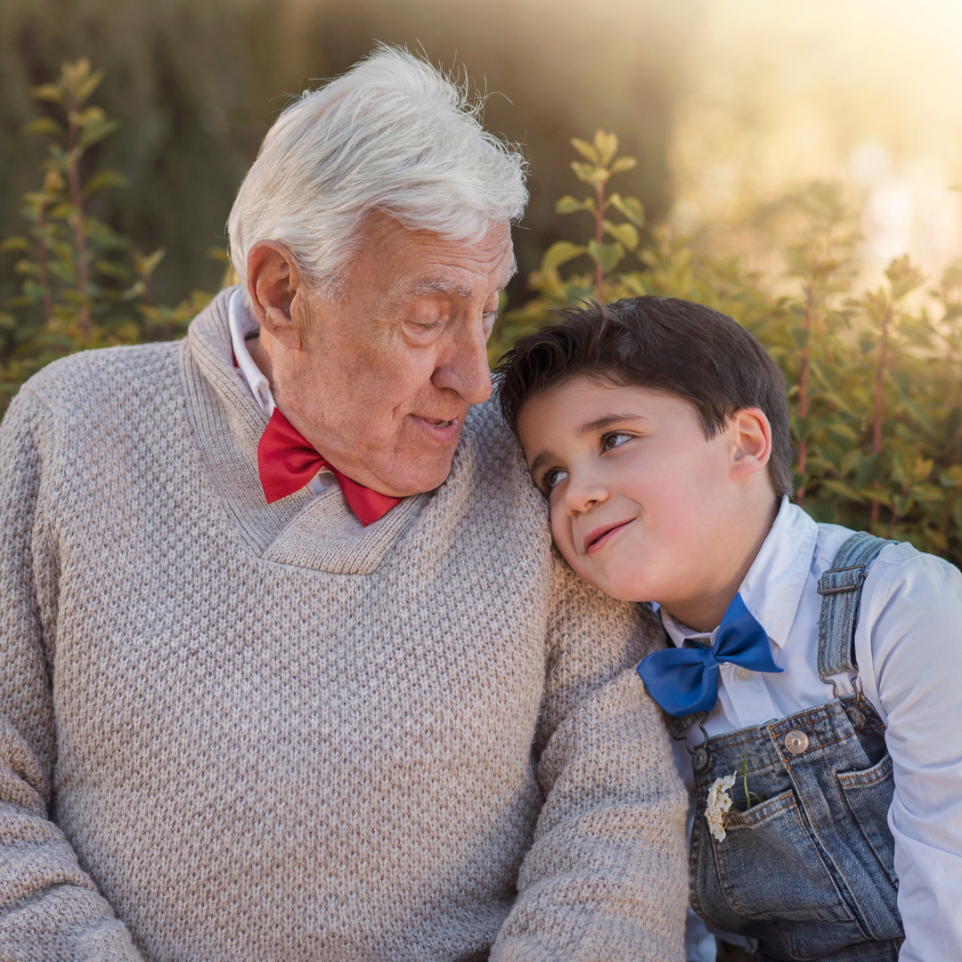 An elderly man is sitting next to a young boy wearing overalls and a bow tie.