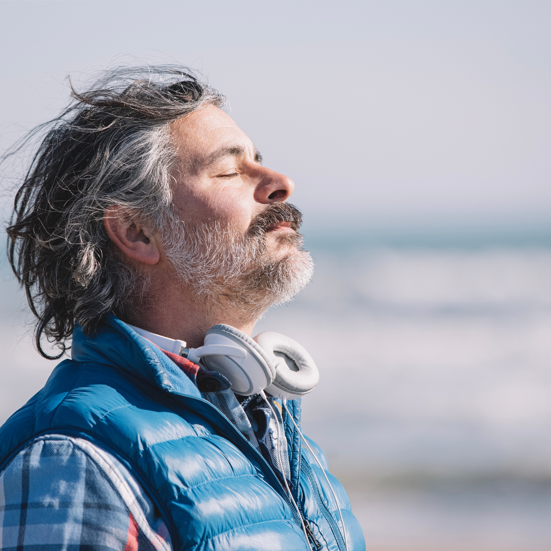 A man wearing headphones is standing on the beach with his eyes closed.