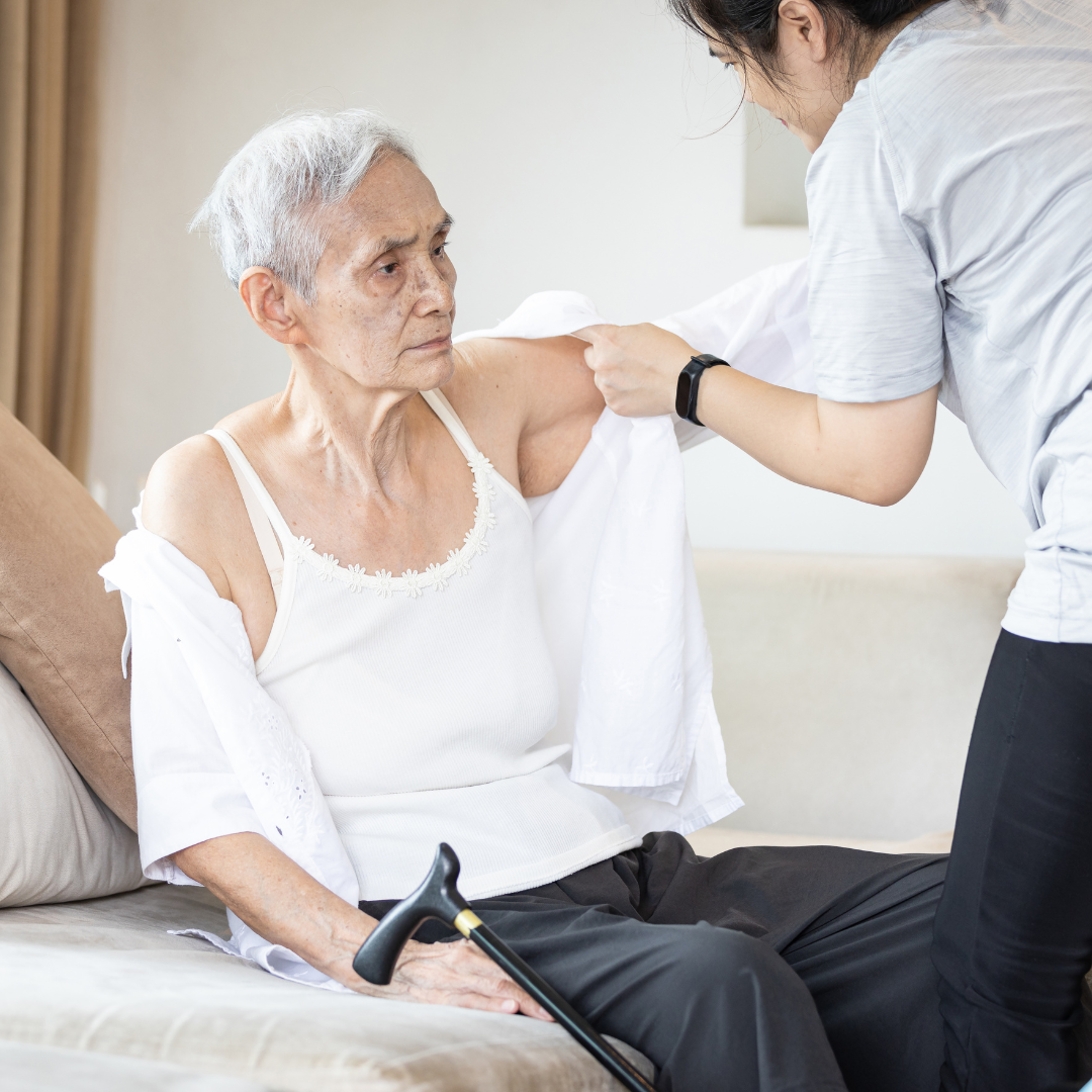 An elderly woman is sitting on a couch with a cane and a nurse is helping her get dressed.