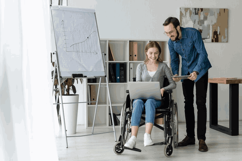 A man is standing next to a woman in a wheelchair using a laptop computer.