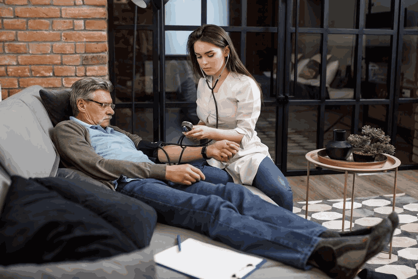 a female caregiver checks the blood pressure of her patient in his home while he relaxes on a couch.