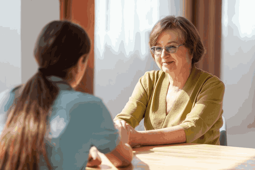 An elderly woman is sitting at a table talking to a younger woman.